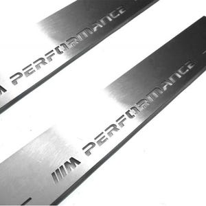 May include: Two silver-colored metal car door sill protectors. The text "M PERFORMANCE" is cut out of the metal. The car accessories are rectangular and have a brushed metal finish. The background is white.