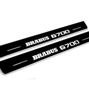 Könnte beinhalten: Zwei schwarze, rechteckige Einstiegsleisten für Autotüren mit dem weißen Text "BRABUS G700". Jeder Schutz hat eine weiße horizontale Linie. Die Artikel werden vor einem weißen Hintergrund angezeigt.
