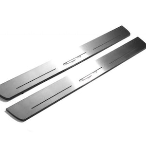 Może przedstawiać: Dwie osłony progów drzwiowych ze szczotkowanego metalu. Każdy prostokątny element ma ciemnoszare wykończenie z dwoma równoległymi rowkami i małym, zakrzywionym wcięciem. Przedmioty są na białym tle.