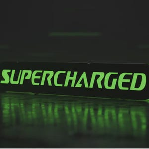 Pode incluir: Uma placa retangular preta com bordas arredondadas, iluminada com a palavra "SUPERCHARGED" em verde brilhante. A placa está sobre uma superfície reflexiva, com um fundo escuro.