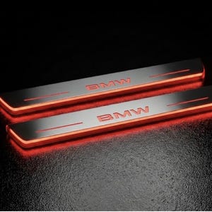 May include: Two illuminated BMW door sill protectors. The silver-colored metal plates have the BMW logo in red. The edges of the plates glow with a bright red light, creating a sleek, modern look.