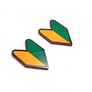 Puede incluir: Dos pegatinas japonesas para conductores principiantes. Cada pegatina es una forma estilizada, verde y amarilla, con un borde negro. Las pegatinas están sobre un fondo blanco.