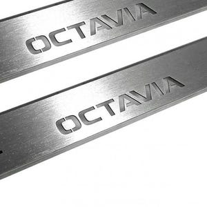 Może przedstawiać: Dwie listwy ochronne progów drzwi samochodu ze szczotkowanego metalu z wygrawerowanym słowem "OCTAVIA" na powierzchni. Metal ma liniowy wzór ziarna. Tło jest białe.