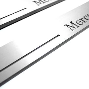 Może przedstawiać: Dwie listwy ochronne progów drzwi samochodu ze szczotkowanego metalu z wygrawerowanymi słowami "Mercedes-Benz" na powierzchni. Ochraniacze mają prostokątny kształt z cienkim rowkiem.