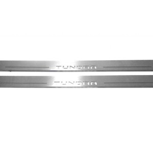 May include: Two silver-colored Tundra door sill protectors. Each protector is rectangular with rounded ends and features the name "TUNDRA" in a bold, sans-serif font. The protectors are designed to be installed on a vehicle.
