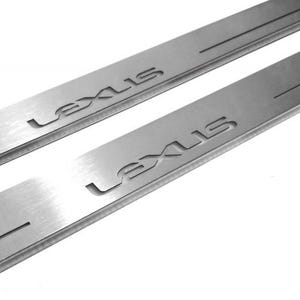 Może przedstawiać: Dwie osłony progów drzwi samochodowych ze szczotkowanego metalu z wygrawerowaną nazwą marki "LEXUS" na powierzchni. Ochraniacze są prostokątne z rowkiem biegnącym wzdłuż. Tło jest białe.