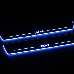 May include: Two black car door sill protectors with bright blue LED lighting. The text "X4" is illuminated in white on each sill. The design features a sleek, modern aesthetic, perfect for automotive customization.