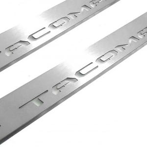 May include: Two brushed metal car door sill protectors. The word "TACOMA" is cut out of the metal. The protectors are rectangular and have a silver finish. The background is white.
