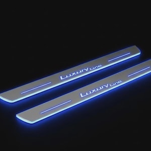 May include: Two illuminated car door sill protectors with the text "Luxury Line" in white. The protectors have a silver finish with a blue neon light strip and text. The background is black.