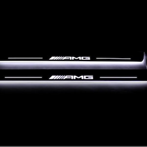 Może przedstawiać: Dwa podświetlane czarne progi drzwi samochodu z białym napisem "AMG". Progi mają biały pasek świetlny wzdłuż dolnej krawędzi i białą linię nad tekstem. Tło jest czarne.