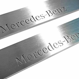 Może przedstawiać: Dwie płyty ze szczotkowanego metalu z wygrawerowanym napisem "Mercedes-Benz". Płyty są prostokątne z zaokrąglonymi narożnikami i szczotkowanym wykończeniem. Tekst jest w kroju bezszeryfowym.
