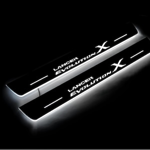 Może przedstawiać: Dwie czarne osłony progów drzwiowych z białym napisem "Lancer Evolution X". Każda osłona ma białą poziomą linię. Elementy są podświetlane białym światłem.