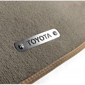 Op de afbeelding: Een beige auto vloermat met een gestikte rand. Een metalen plaat met de naam "TOYOTA" is met twee schroeven aan de mat bevestigd. De mat heeft een getextureerd oppervlak.