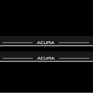May include: Two black car door sill protectors with the brand name "ACURA" in white lettering. Each protector has a white horizontal line. The background is solid black.