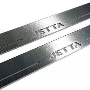 May include: Two brushed metal car door sill protectors with the word "JETTA" etched into the surface. The protectors are rectangular and have a groove cut into the metal. The background is white.