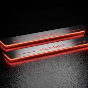 May include: Two illuminated car door sill plates with the word "ALPINA" in silver lettering. The plates have a brushed metal finish and are outlined with a bright red glow. The background is black.
