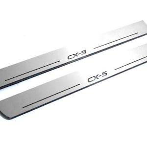 May include: Two silver-colored CX-5 door sill protectors. Each has a black accent line and the CX-5 model name printed in black. The protectors are rectangular with rounded edges, designed for vehicle door entry areas.