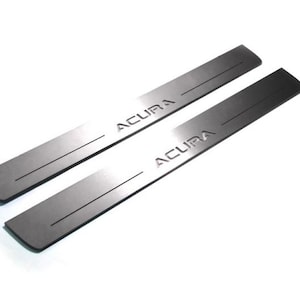 May include: Two silver-colored car door sill protectors. Each has the word "ACURA" engraved in a bold font. The protectors are rectangular with a slightly curved edge and a single groove.