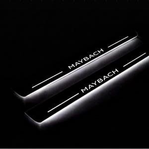 Może przedstawiać: Dwa czarne progi drzwi samochodowych z białym napisem "MAYBACH", podświetlone białym światłem. Progi mają elegancki, prostokątny kształt z cienką białą linią nad tekstem.