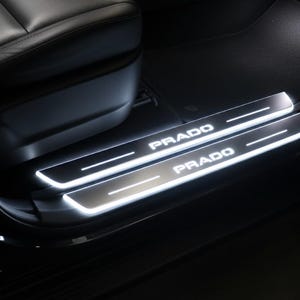 May include: Illuminated silver door sills with the name "PRADO" in white lettering. The sills are installed in a vehicle, near the front seat. The sills have a bright, white glow.