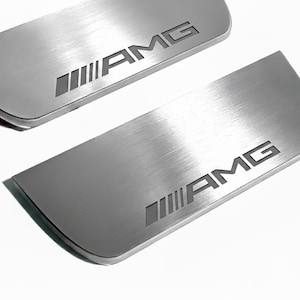 May include: Two brushed metal car door sill plates with the AMG logo. The plates are silver with a curved edge and the AMG logo is etched in a darker gray. The background is white.