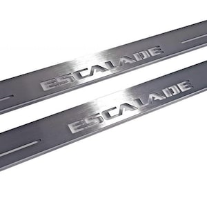 May include: Two brushed metal door sill plates with the word "ESCALADE" embossed on each. The plates have a linear groove and are angled towards the top right. The background is white.