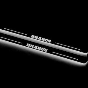 Może przedstawiać: Dwie podświetlane listwy progowe z nazwą marki "BRABUS" w kolorze białym, na czarnym tle. Listwy mają elegancki, nowoczesny design z efektem świecenia.