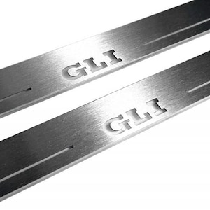 May include: Two brushed metal car door sill protectors. Each has the word "GLI" cut out of the metal. A thin groove runs across each protector. The background is white.
