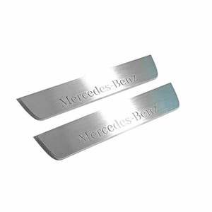 May include: Two brushed metal car door sill protectors. Each has a rectangular shape with rounded corners and the engraved text "Mercedes-Benz". The protectors are silver and are set against a white background.