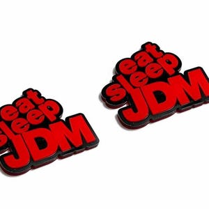 Può includere: Due adesivi JDM rossi e neri con le parole "eat sleep JDM" in un design impilato. Gli adesivi hanno una finitura lucida e sono su uno sfondo bianco.