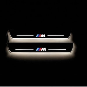 May include: Two black car door sills with the BMW M logo in blue, red, and white. Each sill has a white horizontal light and the M logo. The sills are illuminated against a dark background.