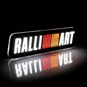Könnte beinhalten: Beleuchtetes schwarzes rechteckiges Schild mit dem Wort "RALLI ART" in Weiß, mit einem rot-orangen Streifendesign. Das Schild spiegelt sich auf einer dunklen Oberfläche.