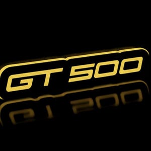 May include: A three-dimensional, gold-colored "GT 500" emblem against a black background. The letters and numbers are outlined in gold, with a slight shadow effect. The emblem's reflection is visible below.