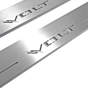 May include: Two brushed metal car door sill protectors. Each has the word "COLT" cut out in a blocky font, with a stylized emblem below. The metal has a matte finish and a few thin cut-out lines.