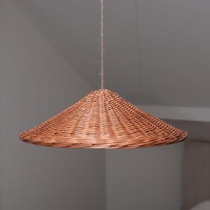 May include: A woven, conical pendant light fixture. The light is made of light brown wicker and hangs from a natural fiber cord. The shade has a wide, flared shape, and the weave pattern is visible.