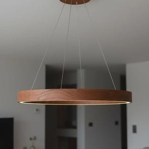 May include: A contemporary circular pendant light. The light has a dark wood ring with a warm, diffused light source. It is suspended from the ceiling by thin wires, and the ceiling mount is also made of wood.