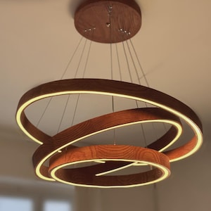 May include: A circular pendant light fixture featuring three interconnected rings with a wood grain finish. Each ring contains an integrated LED light strip that emits a warm glow. The fixture is suspended from a matching circular ceiling mount.