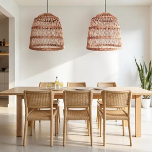 Op de afbeelding: Een eethoek met een lichte houten tafel en zes geweven stoelen. Twee geweven hanglampen hangen boven de tafel. Een groot raam en een plant in een pot zijn zichtbaar. De kamer heeft een neutraal kleurenpalet.