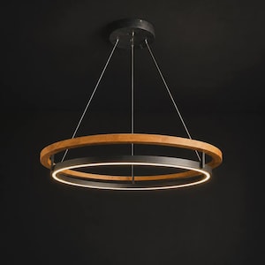 May include: A circular pendant light fitting featuring a natural wood outer ring and a matte black inner ring. The inner ring contains an integrated LED light strip that emits a soft glow. The fitting is suspended by thin metal cables.