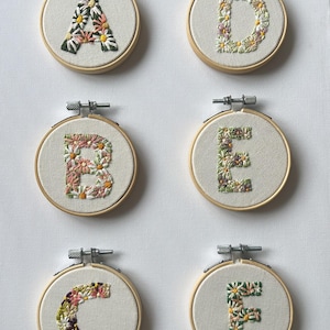May include: Six circular wooden embroidery hoops containing off-white fabric panels. Each hoop features a hand-stitched letter of the alphabet, A through F, intricately filled with colourful floral patterns in shades of pink, yellow, white, and green.