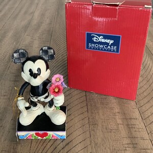Op de afbeelding: Een Mickey Mouse-figuurtje dat een boeket roze en paarse bloemen vasthoudt, staand op een rechthoekige voet met een rood hartontwerp. Daarnaast staat een rode Disney Showcase Collection-doos met witte tekst en een blauw logo op de voorzijde.
