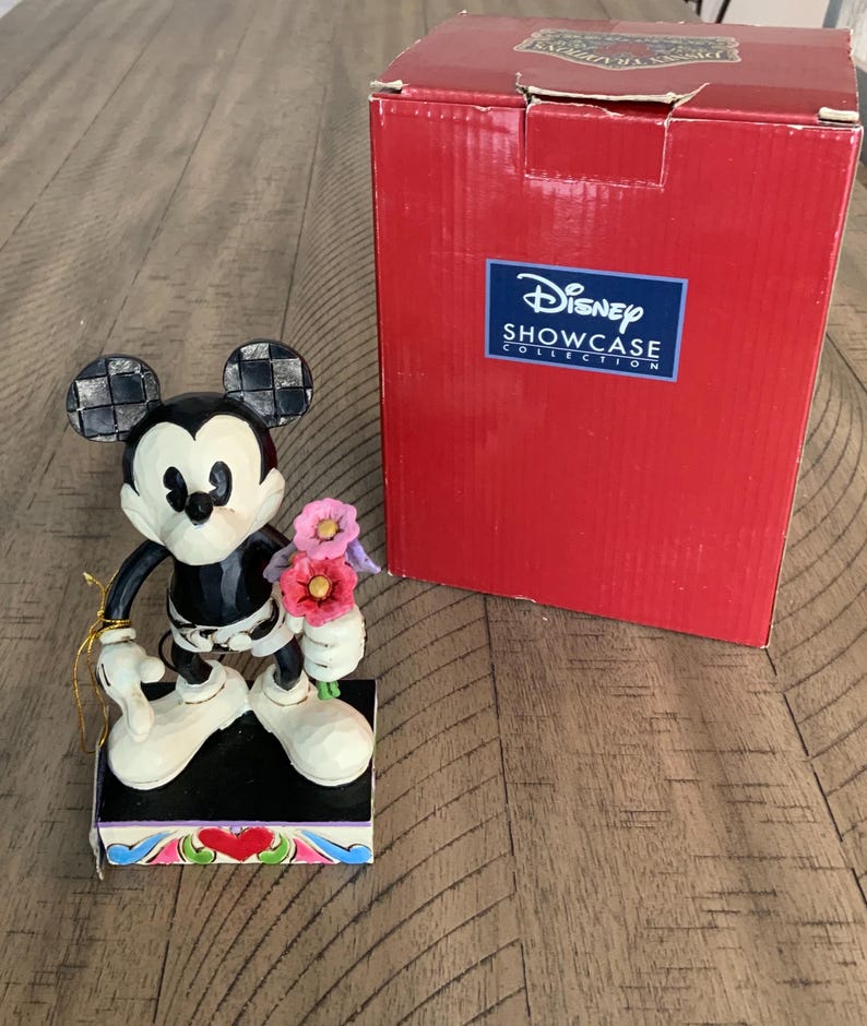 Op de afbeelding: Een Mickey Mouse-figuurtje dat een boeket roze en paarse bloemen vasthoudt, staand op een rechthoekige voet met een rood hartontwerp. Daarnaast staat een rode Disney Showcase Collection-doos met witte tekst en een blauw logo op de voorzijde.
