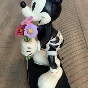 Op de afbeelding: Een Mickey Mouse-beeldje met een zwart-wit kleurenschema en een uitgesneden textuur. Het personage zit op de knie&euml;n en houdt een klein boeket van rode, roze en paarse bloemen vast. Het figuur staat op een rechthoekige basis.