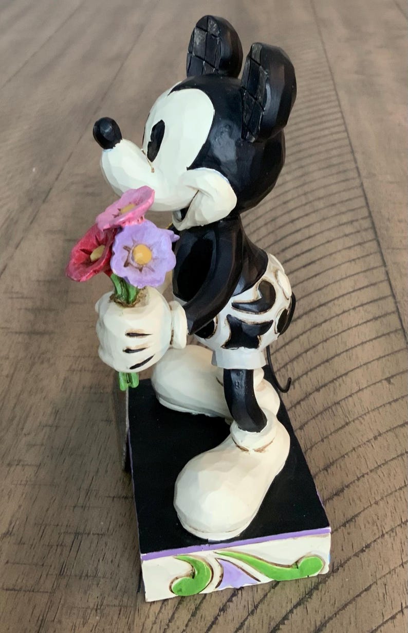 Op de afbeelding: Een Mickey Mouse-beeldje met een zwart-wit kleurenschema en een uitgesneden textuur. Het personage zit op de knie&euml;n en houdt een klein boeket van rode, roze en paarse bloemen vast. Het figuur staat op een rechthoekige basis.