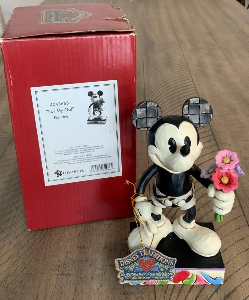 Op de afbeelding: Een Mickey Mouse-figuurtje dat een boeket roze, paarse en rode bloemen vasthoudt, staand op een zwarte voet met een Disney Traditions-label. Daarnaast staat een rode kartonnen doos met het label 4043665 For My Gal Figurine met een afbeelding.