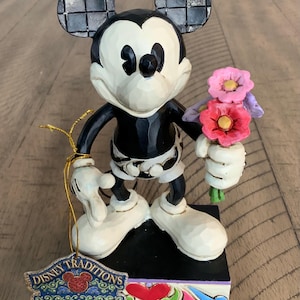 Op de afbeelding: Een Mickey Mouse-figuur met een zwart-wit lichaam, grote oren met een zwart ruitpatroon en witte schoenen. Het personage houdt een boeket roze en rode bloemen vast. Een Disney Traditions Jim Shore Enesco-label is bevestigd met een gouden koord.