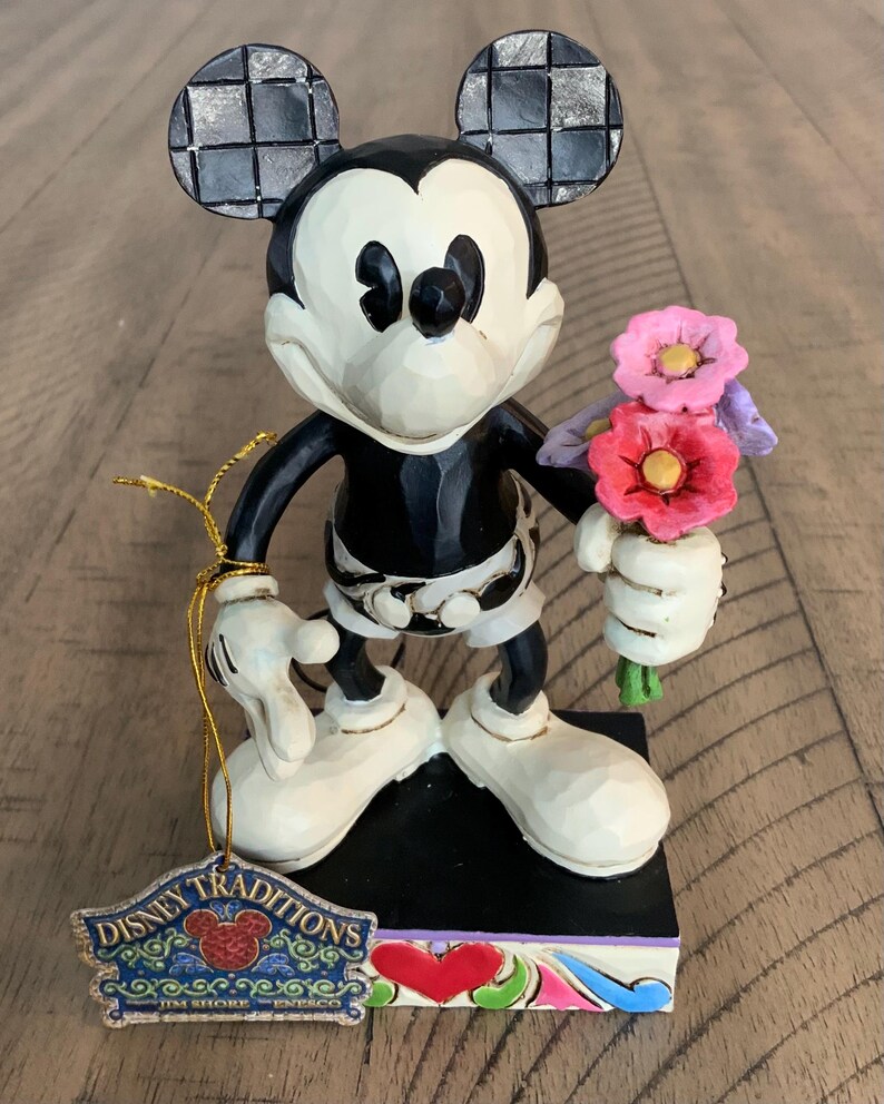 Op de afbeelding: Een Mickey Mouse-figuur met een zwart-wit lichaam, grote oren met een zwart ruitpatroon en witte schoenen. Het personage houdt een boeket roze en rode bloemen vast. Een Disney Traditions Jim Shore Enesco-label is bevestigd met een gouden koord.