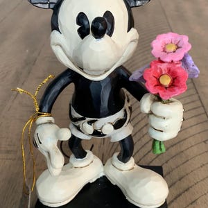 Op de afbeelding: Een Mickey Mouse-figuur in zwart-wit met geblokte oren, die een klein boeket roze en paarse bloemen vasthoudt. Het personage staat op een rechthoekige voet die is versierd met een rood hart en kleurrijke krulpatronen.