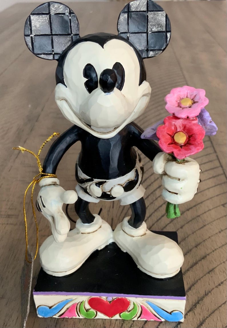 Op de afbeelding: Een Mickey Mouse-figuur in zwart-wit met geblokte oren, die een klein boeket roze en paarse bloemen vasthoudt. Het personage staat op een rechthoekige voet die is versierd met een rood hart en kleurrijke krulpatronen.