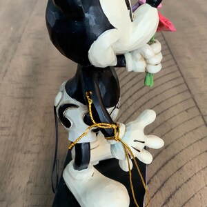 Op de afbeelding: Een Mickey Mouse-figuur met een uitstraling van gesneden hout, geschilderd in zwart, wit en cr&egrave;me. Het personage houdt een klein boeket van roze en rode bloemen vast. Een dunne gouden metalen draad is om de arm en taille gewikkeld.